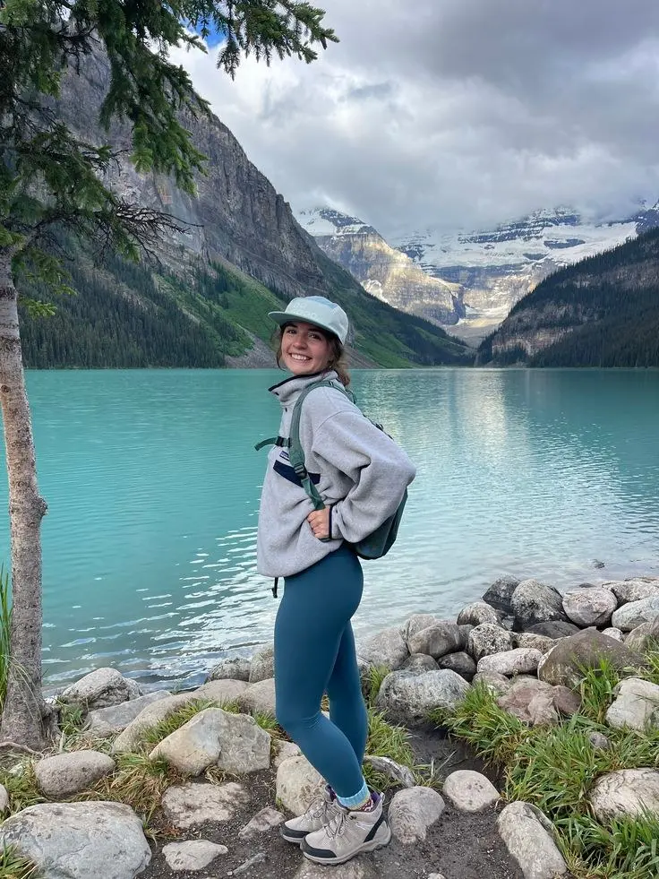 woman in grey fleece zip pullover teal blue leggings tan hiking boots small backpack snapback hat smiling on rocky lakeside trail photo 9
