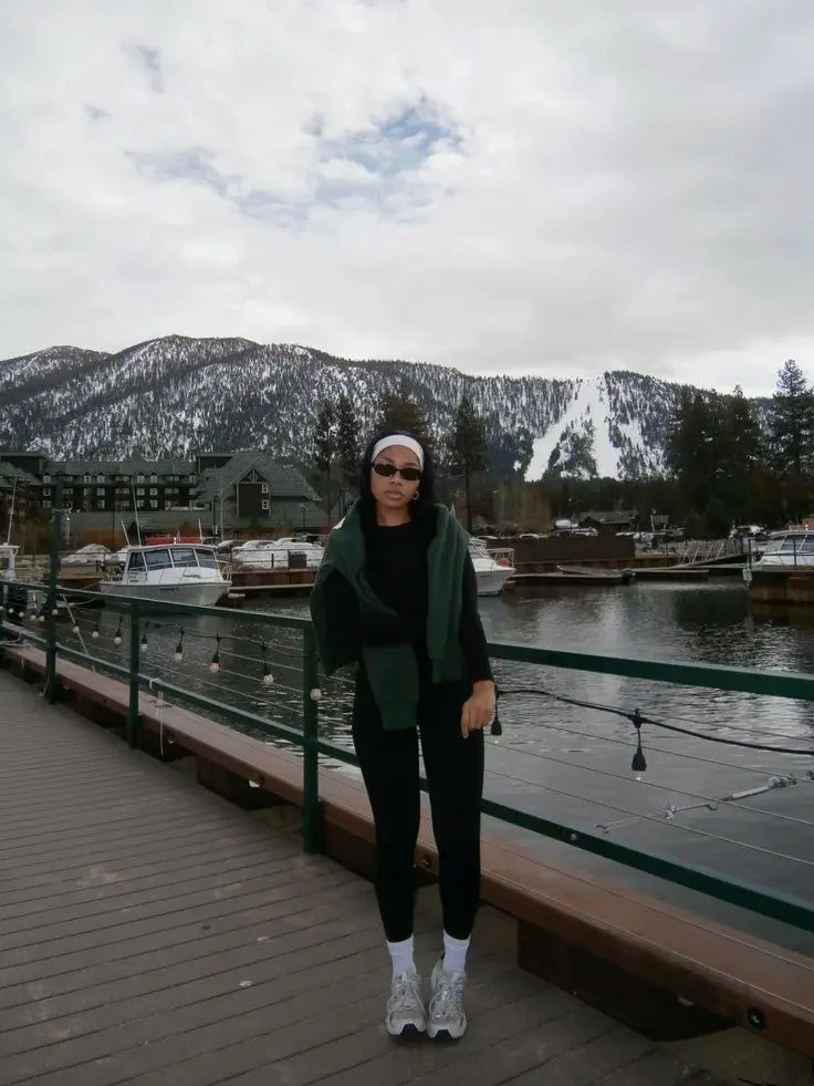 woman in black leggings black long sleeve base layer green fleece tied at waist white sports headband small sunglasses silver trail sneakers on waterfront boardwalk with snowy mountains photo 10