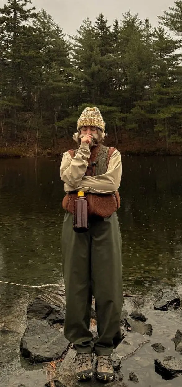 woman in cream long sleeve base layer tan fleece zip sherpa brown vest wide olive waterproof pants hiking boots speckled knit beanie holding thermos standing on rocks by lake in rain photo 12