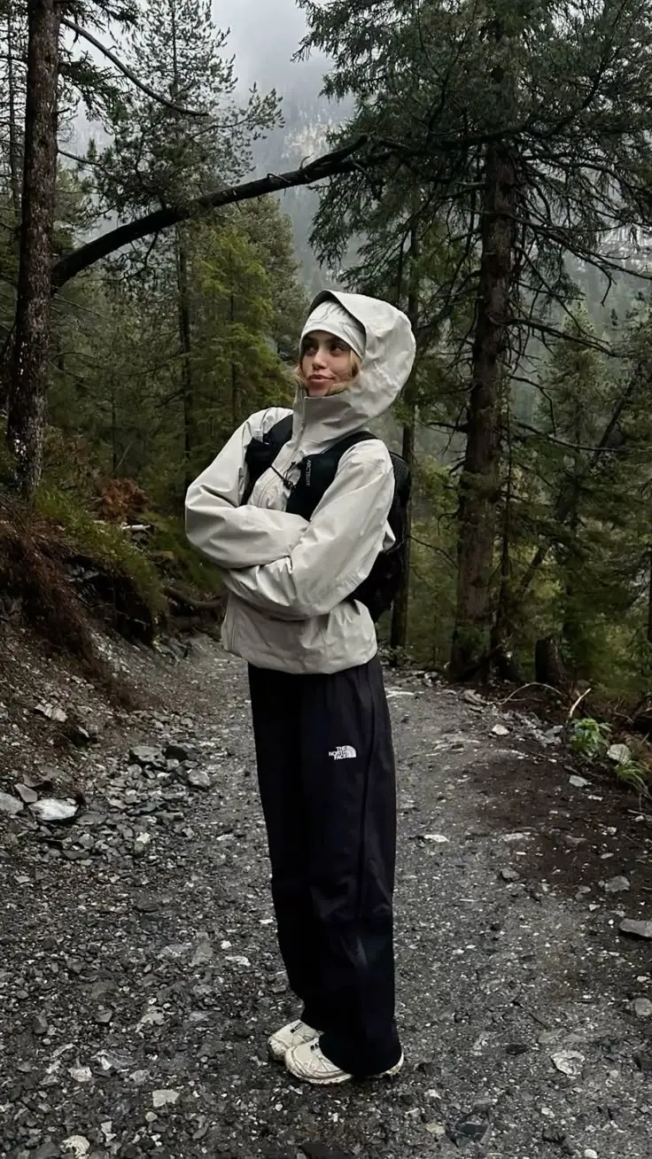 woman in cream hooded shell jacket hood up wide black technical pants white trail sneakers standing on rocky mountain path in rain photo 15