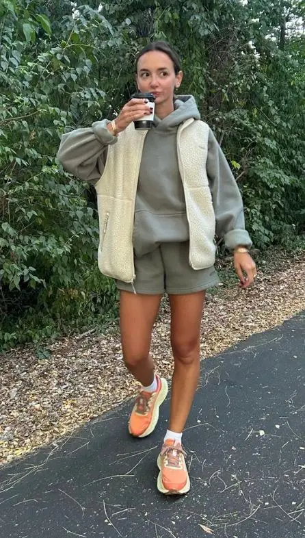 woman in sage green hoodie cream sherpa jacket olive sweat shorts bright orange green sneakers white socks walking on paved path holding coffee cup photo 16