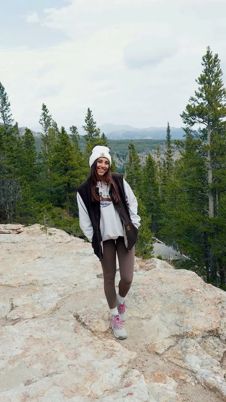 woman in white pom beanie brown canvas duck vest grey graphic sweatshirt brown taupe leggings grey hiking shoes pink laces white socks smiling on rocky mountain summit photo 19