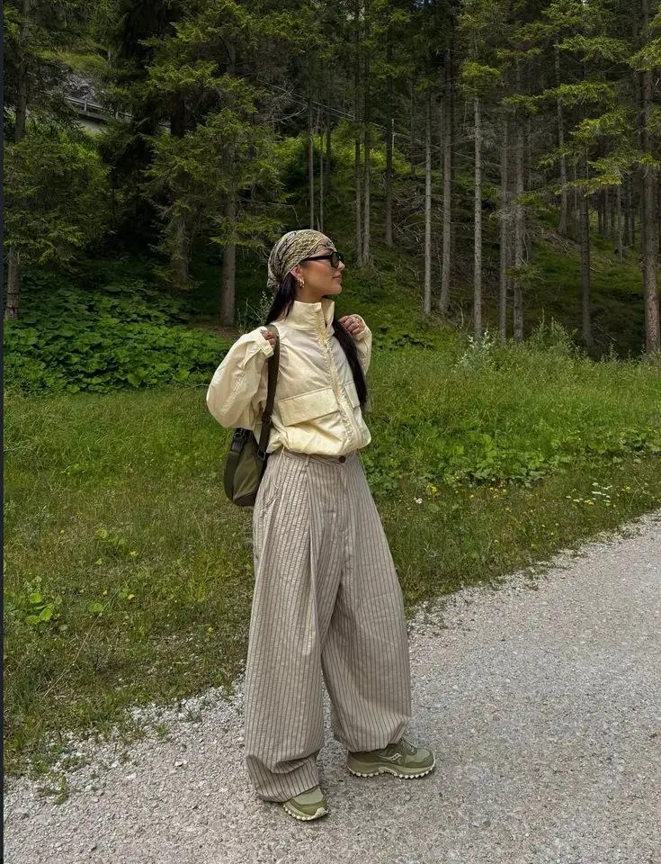 woman in pale yellow cropped puffer jacket wide pinstripe cream trousers olive trail sneakers printed silk bandana headscarf small sunglasses olive backpack on gravel forest path photo 20