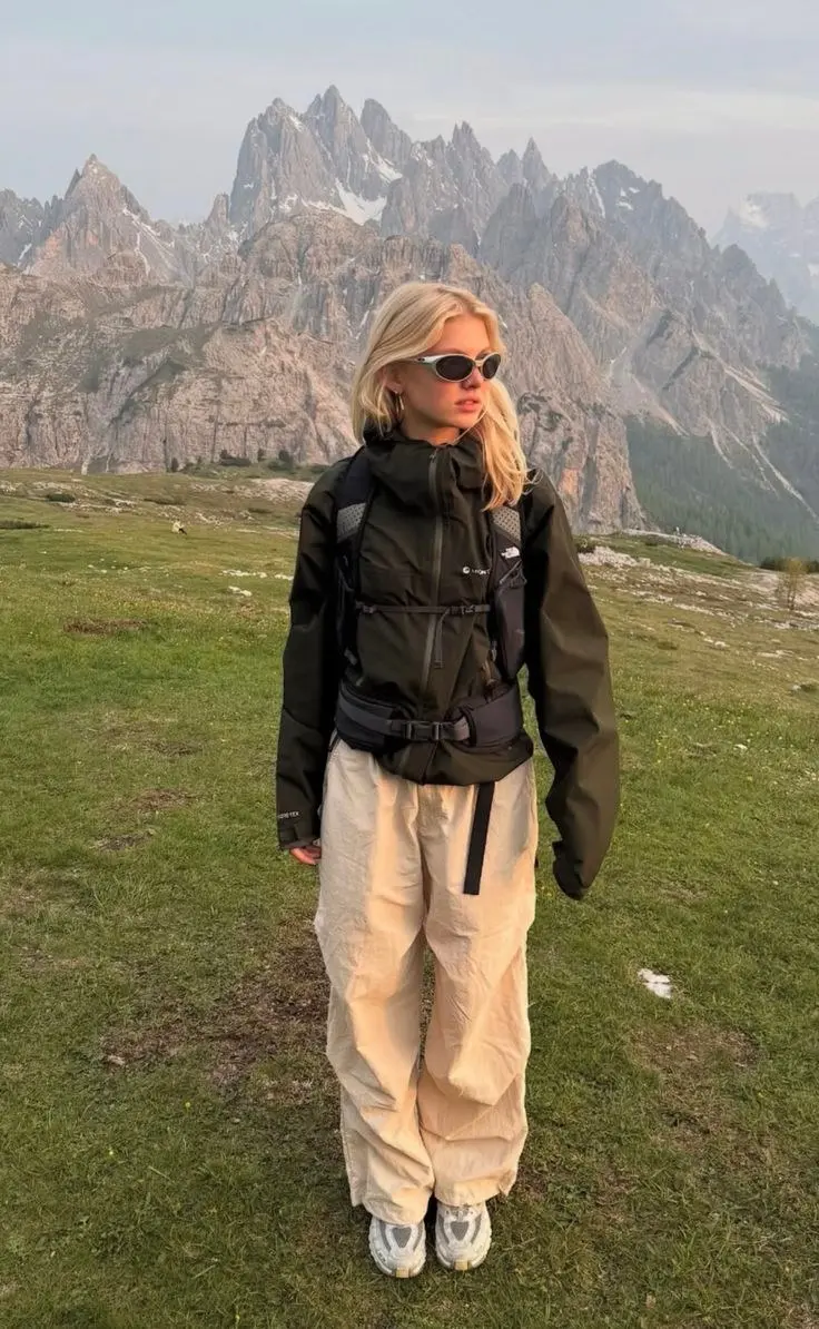 woman in black hooded Gore-Tex shell jacket cream wide parachute pants large technical hiking backpack white oval sunglasses trail runners standing on alpine meadow with dramatic mountain peaks photo 22