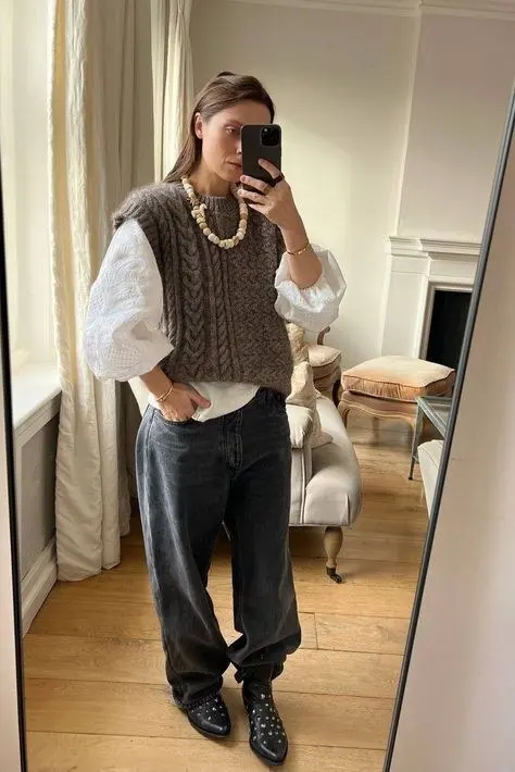 woman wearing brown cable knit sweater vest white puff sleeve blouse dark grey jeans studded black ankle boots wooden bead necklace mirror selfie outfit 2