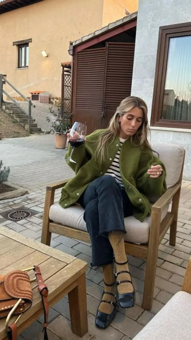woman seated outdoors wearing oversized olive green cardigan coat navy striped tee dark raw hem jeans buckled mary jane shoes sheer socks leather bag on table outfit 4