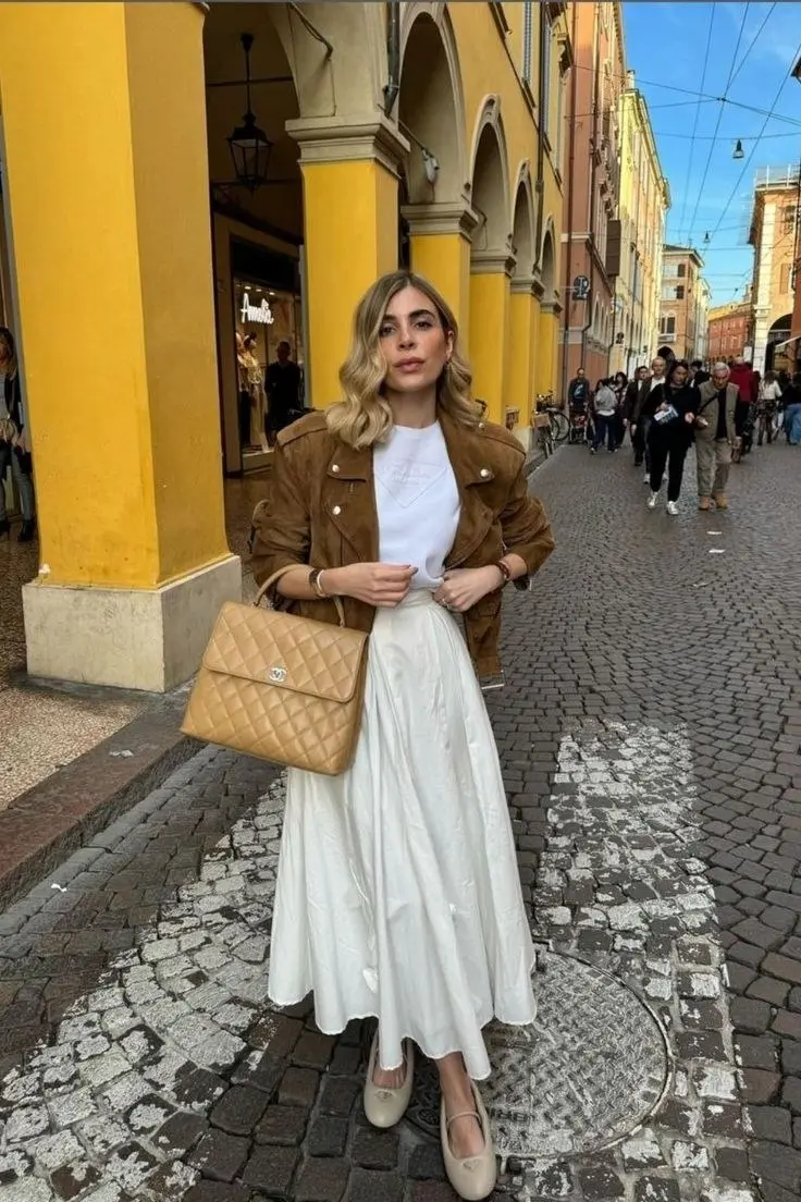 woman wearing tan suede moto jacket white graphic tee flowing white midi skirt camel quilted chain bag ivory ballet flats cobblestone street outfit 5