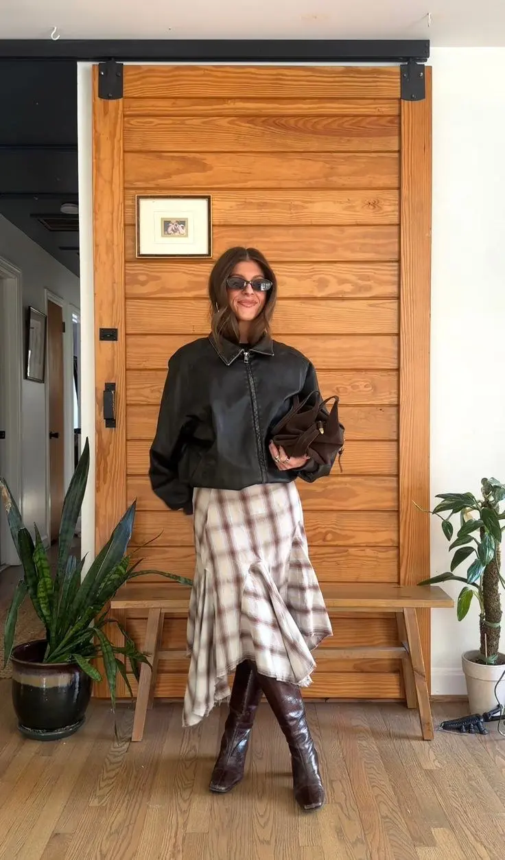 woman wearing oversized black leather bomber jacket cream caramel plaid bias cut midi skirt dark brown knee high boots dark chocolate hobo bag small grey lens sunglasses half up hair indoor outfit 11