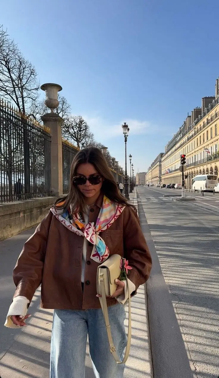 woman wearing cognac brown leather overshirt jacket white crewneck wide leg light wash jeans multicolor printed silk neck scarf cream mini shoulder bag small flower detail sunglasses outdoor street outfit 12