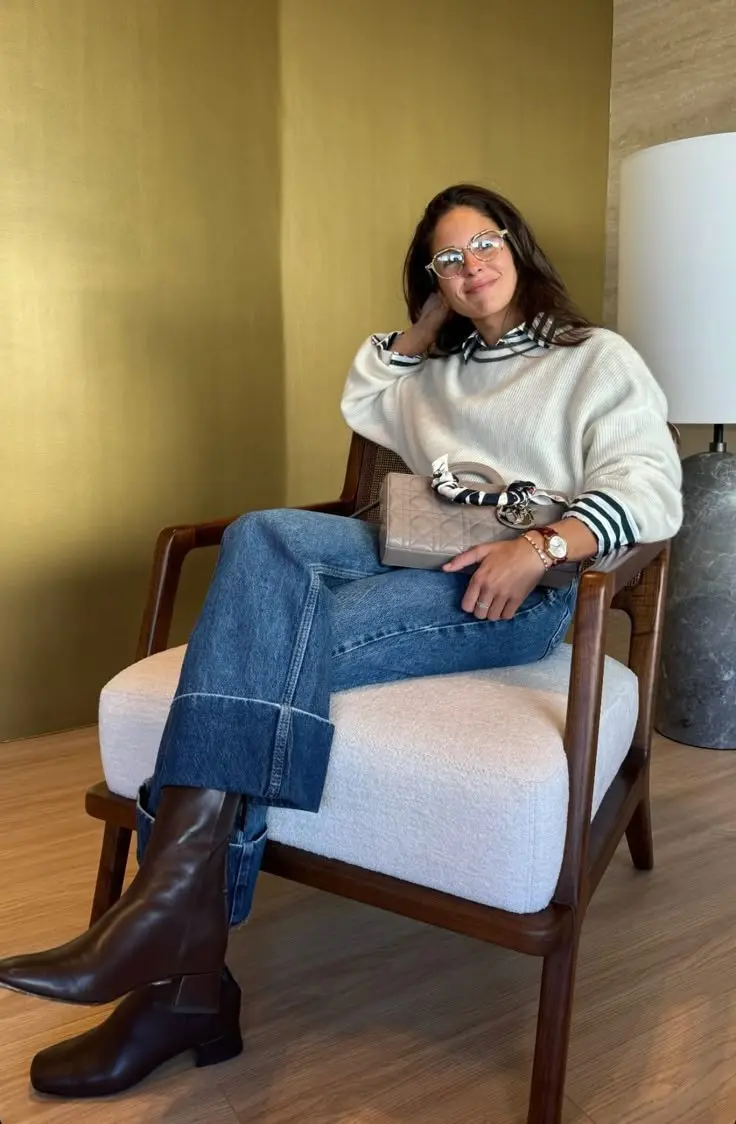 woman wearing cream ribbed crewneck sweater navy striped shirt layered collar and cuffs wide leg medium wash jeans dark brown knee high boots round clear frame glasses quilted bag seated chair outfit 13