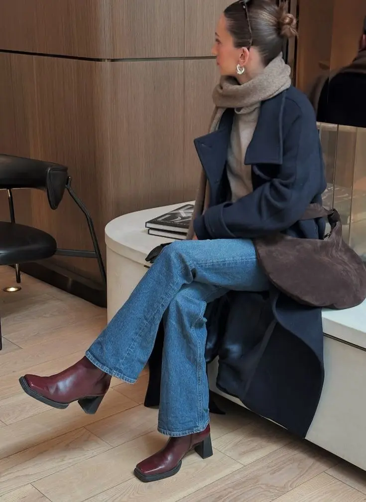 woman seated wearing long navy structured coat camel turtleneck wide leg blue jeans burgundy block heel square toe boots chocolate suede crescent bag gold hoop earrings indoor lobby outfit 14