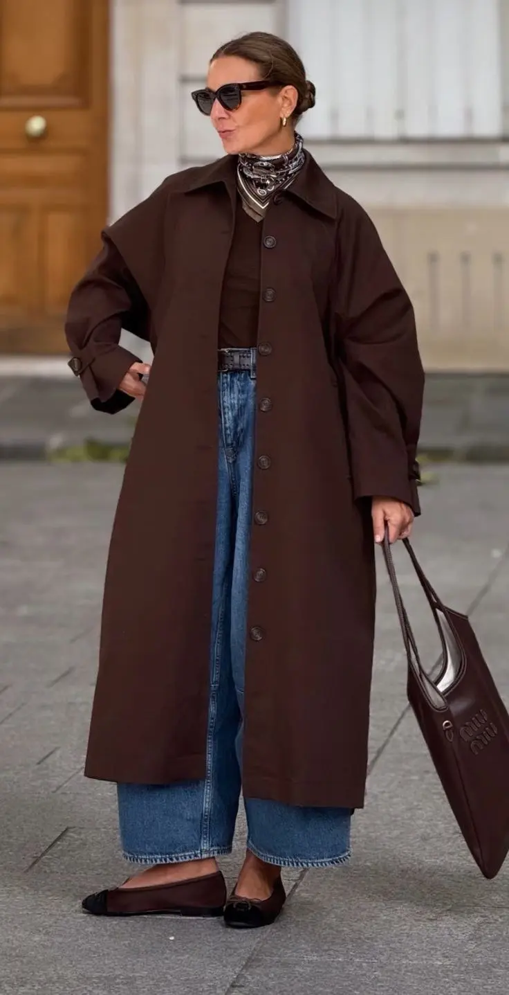 woman standing street wearing long deep chocolate brown button front trench coat open medium wash wide leg jeans dark ballet flats printed silk neck scarf dark oversized sunglasses elongated leather bag updo hair outfit 19