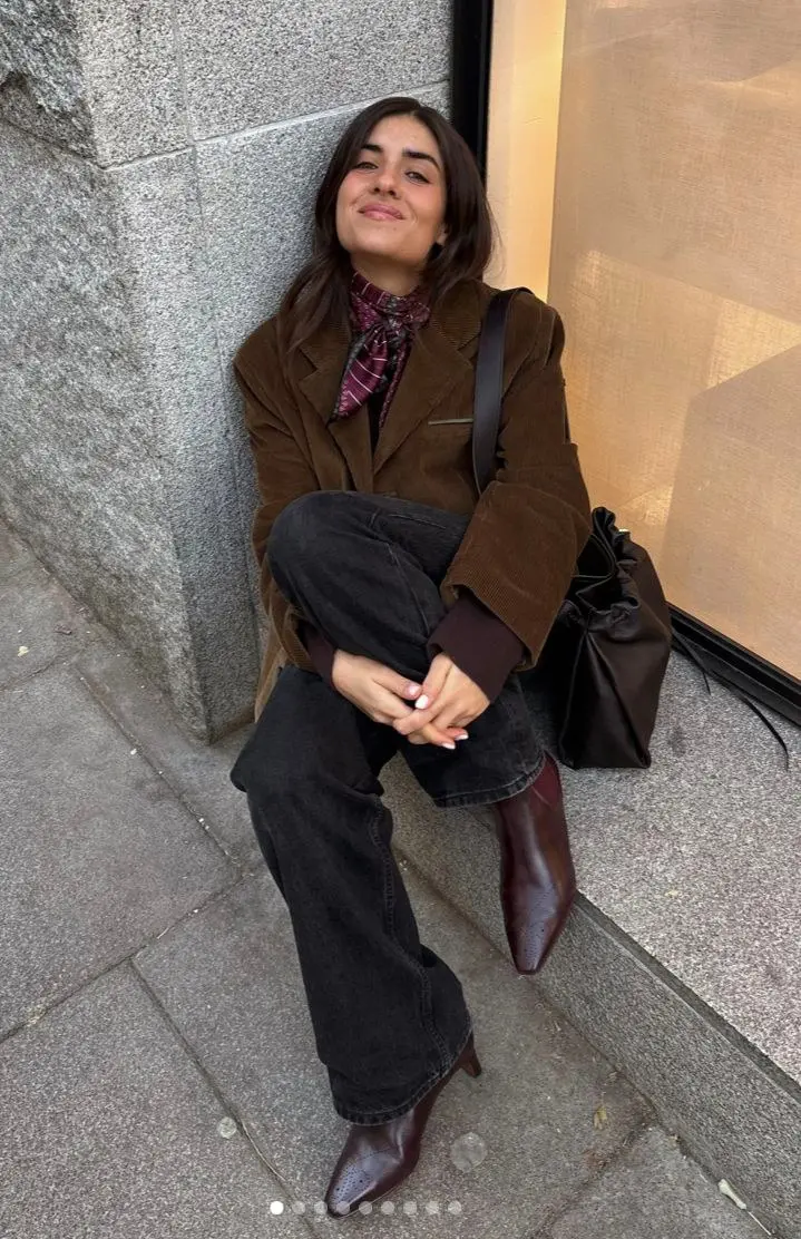 woman seated outdoors wearing chocolate brown corduroy blazer burgundy collar shirt printed neckerchief scarf black wide leg jeans dark brown knee high pointed boots dark drawstring shoulder bag smiling outdoor granite wall outfit 20