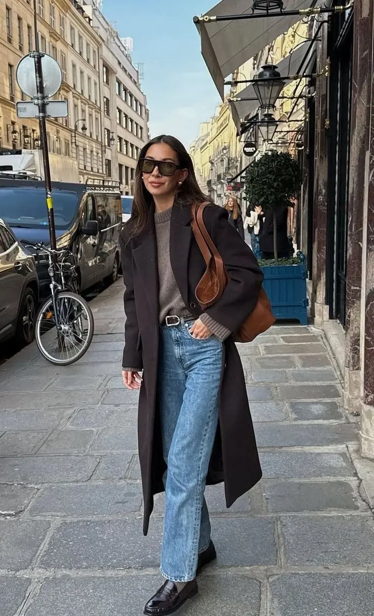 woman walking street wearing long dark charcoal brown duster coat open taupe grey crewneck sweater medium wash straight jeans dark burgundy penny loafers large cognac leather tote bag pearl stud earrings sunglasses outfit 22