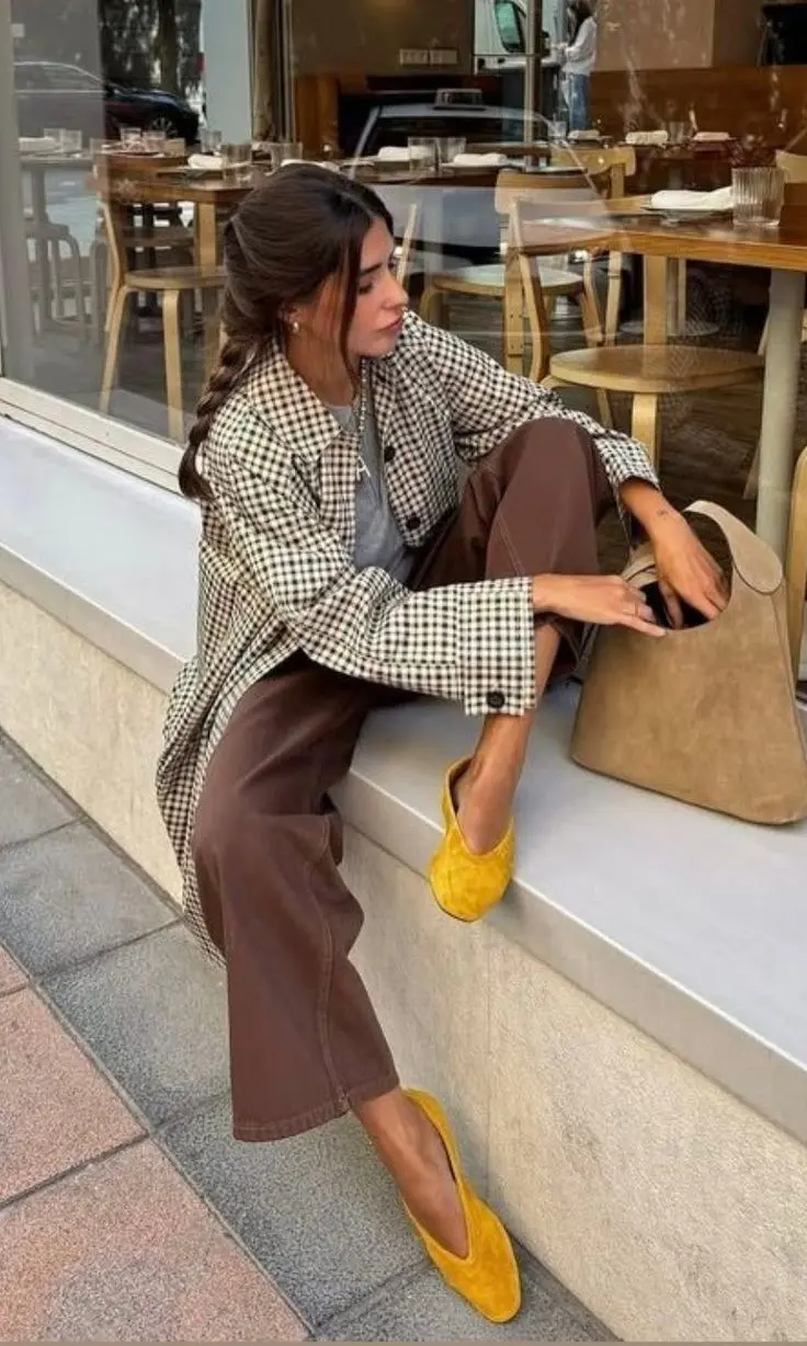 woman seated outdoor cafe wearing oversized black cream gingham blazer grey tee warm brown wide leg trousers bright yellow suede ballet flats small sand suede tote bag side braid updo pearl drop earrings outfit 23