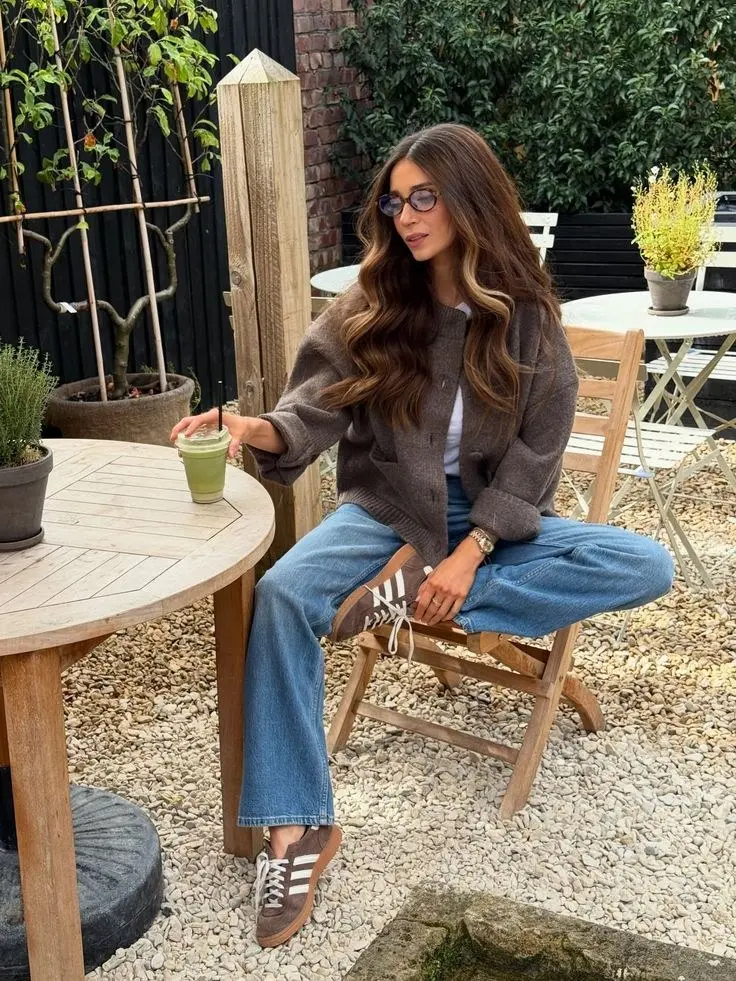 woman seated outdoor garden cafe wearing taupe brown zip front knit cardigan white tee medium wash wide leg jeans brown white adidas gazelle sneakers round lavender tinted glasses watch ring green juice table outfit 24