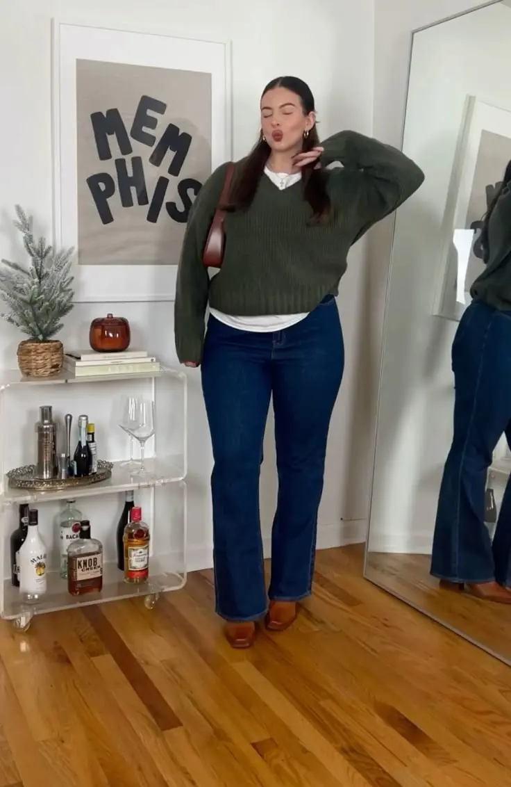 curvy woman in forest green vneck sweater white shirt layered underneath dark flare jeans tan block heel boots brown crossbody bag indoor outfit 10