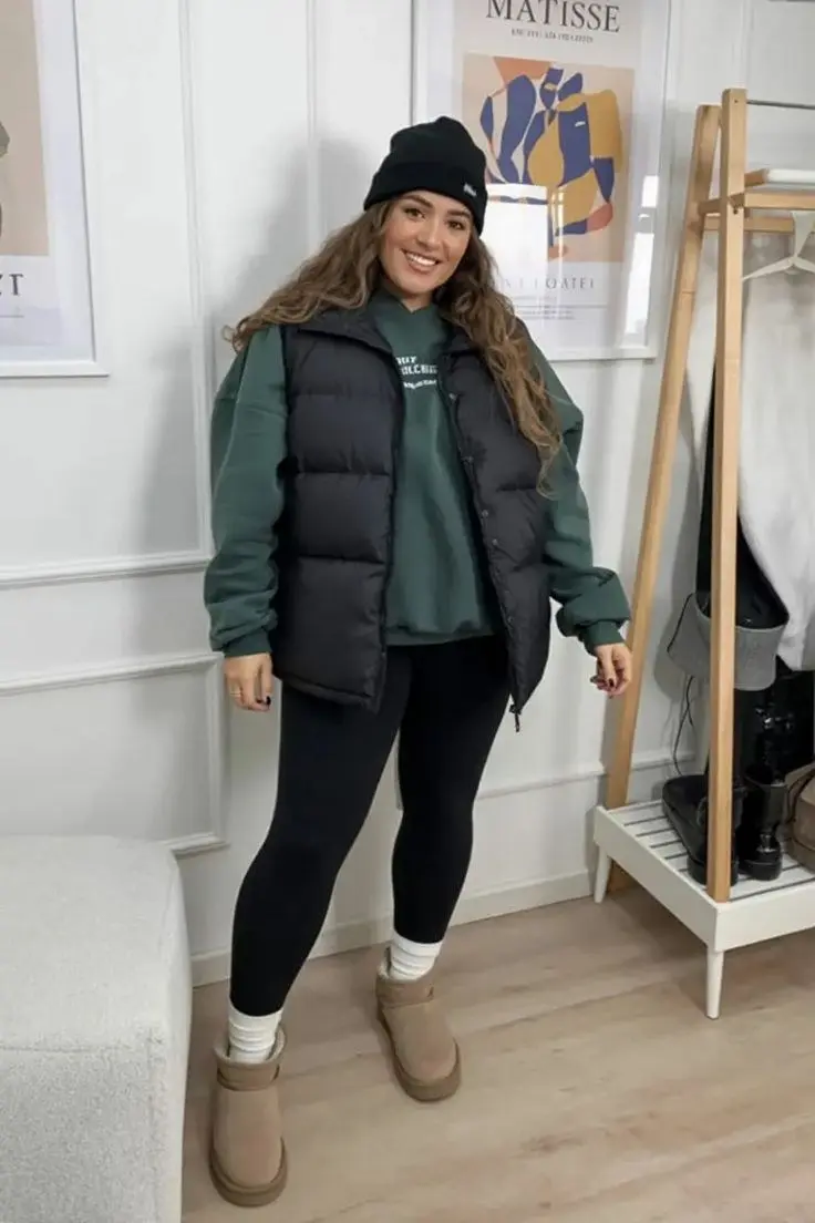 curvy woman in forest green hoodie black puffer vest black leggings white cuffed platform boots black beanie indoor casual outfit 11