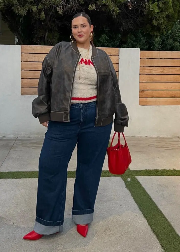 curvy woman in distressed leather bomber jacket ivory cable knit sweater cuffed wide leg dark jeans red pointed mules red structured tote gold hoop earrings outdoor outfit 12