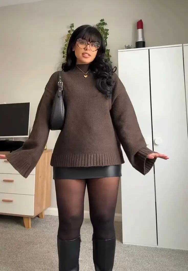curvy woman in brown oversized flare sleeve chunky sweater black faux leather mini skirt sheer black tights knee high black boots small black saddle bag tortoiseshell glasses heart pendant necklace mirror selfie outfit 20