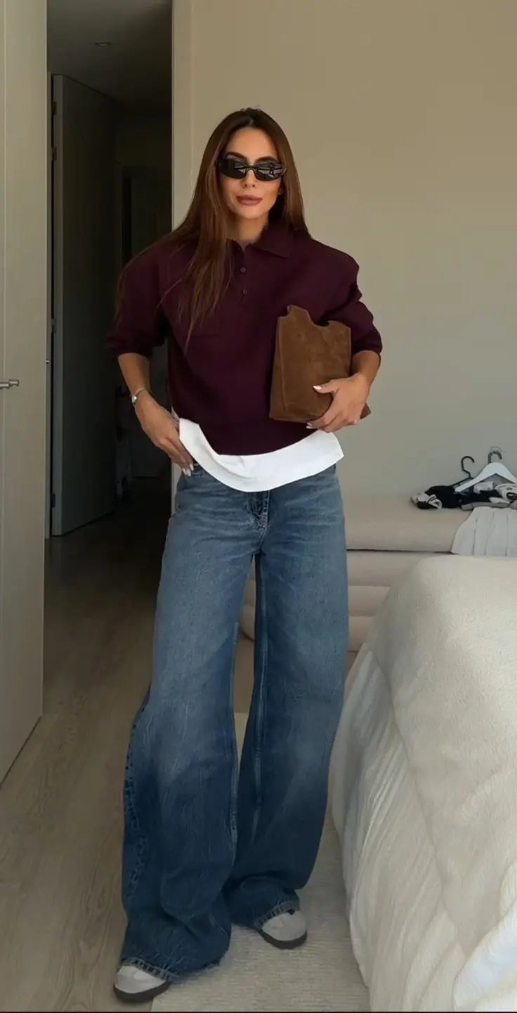 woman in burgundy polo knit sweater layered over white shirt medium blue wide leg jeans grey sneakers tan suede clutch sunglasses mirror outfit 4