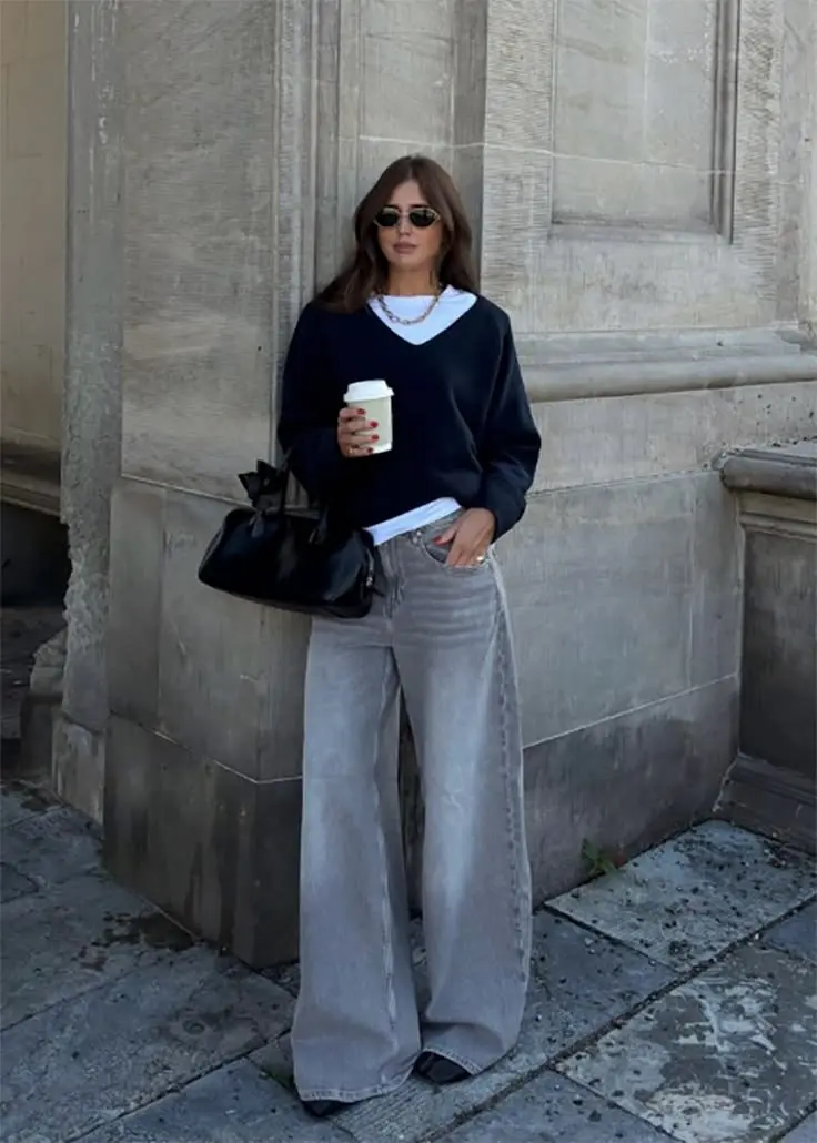 woman in black v-neck knit sweater layered over white shirt light grey wide leg jeans black kitten heel shoes gold chain necklace black soft bag coffee cup street outfit 16