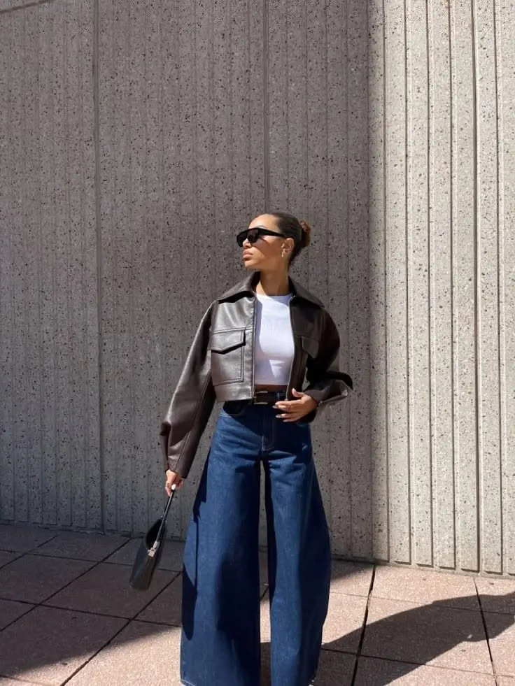 woman in cropped dark brown leather jacket white crop tee black belt dark blue wide leg jeans bun hairstyle sunglasses slim black bag outdoor outfit 21