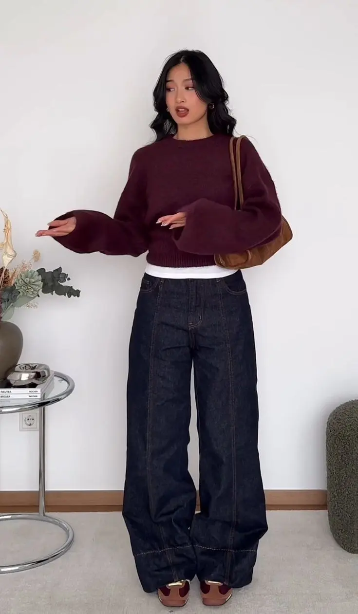 woman in burgundy oversized crewneck knit white underlayer dark navy wide leg jeans gold loafer mules tan suede shoulder bag sunglasses indoor bedroom outfit 23