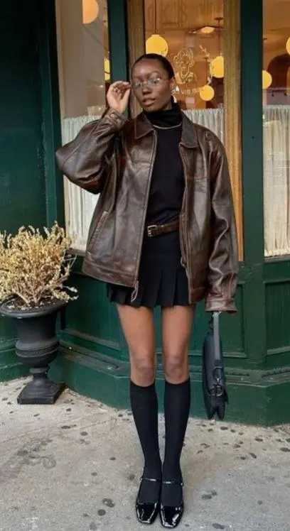 woman in oversized distressed brown leather jacket black turtleneck black pleated mini skirt knee socks patent Mary Janes black shoulder bag street style outfit 2