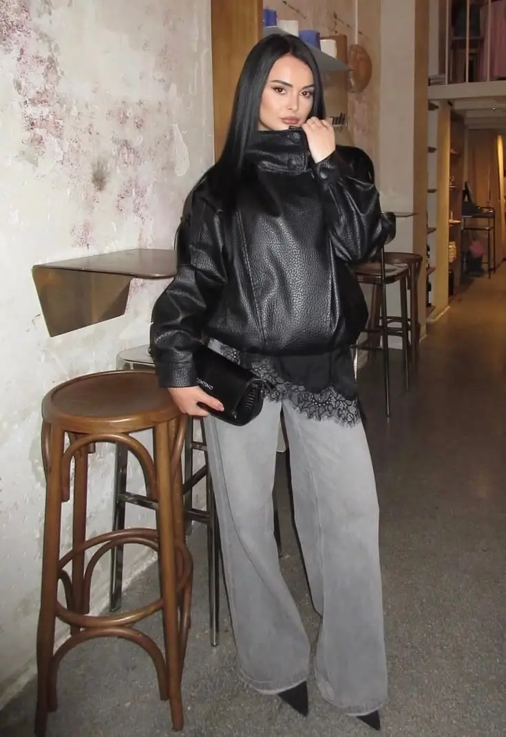 woman in oversized black leather jacket with shearling collar lace trim top grey wide leg jeans black pointed heeled boots black clutch bag cafe interior outfit 4