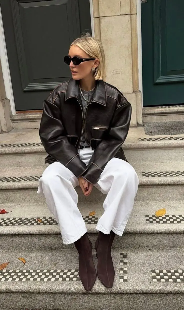 woman sitting on steps in dark brown oversized leather bomber jacket grey henley white wide leg jeans dark brown pointed ankle boots silver jewelry narrow oval sunglasses outdoor outfit 11
