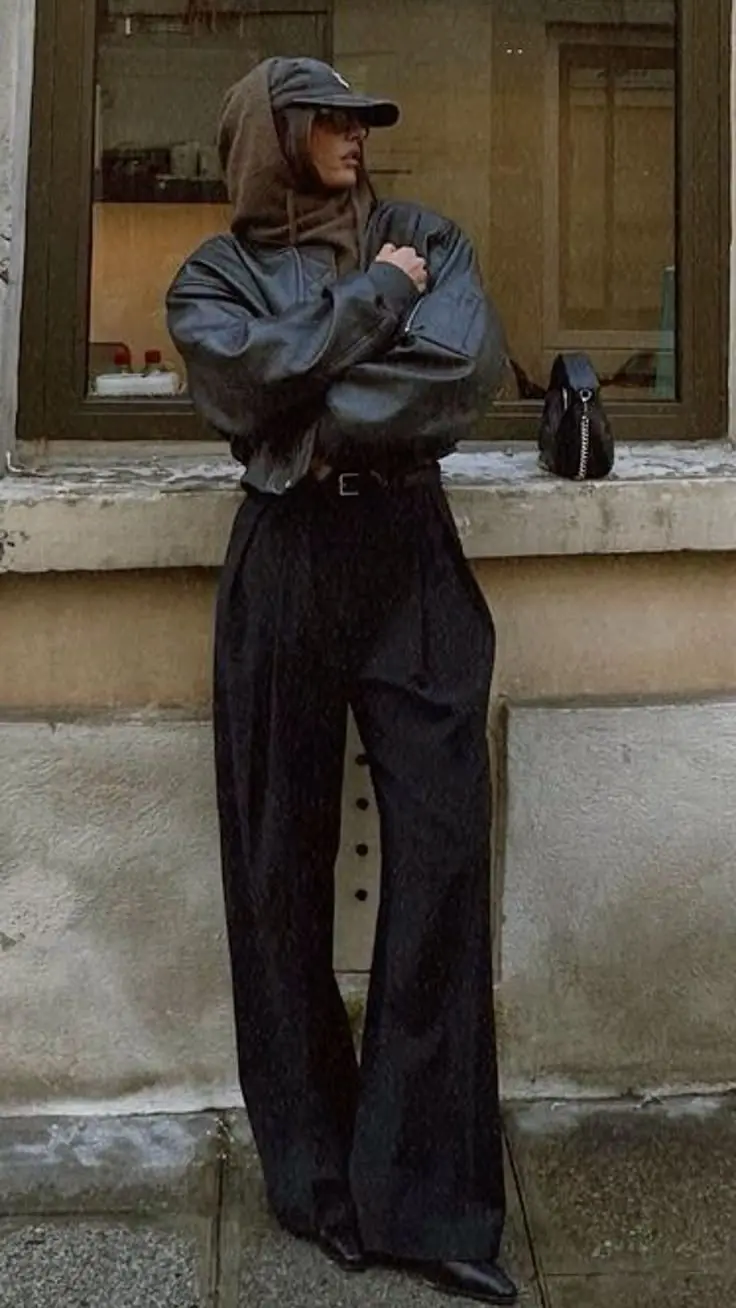 woman in dark leather bomber jacket brown hoodie cap black wide leg tailored trousers black belt black pointed boots small black chain bag leaning against wall outfit 12