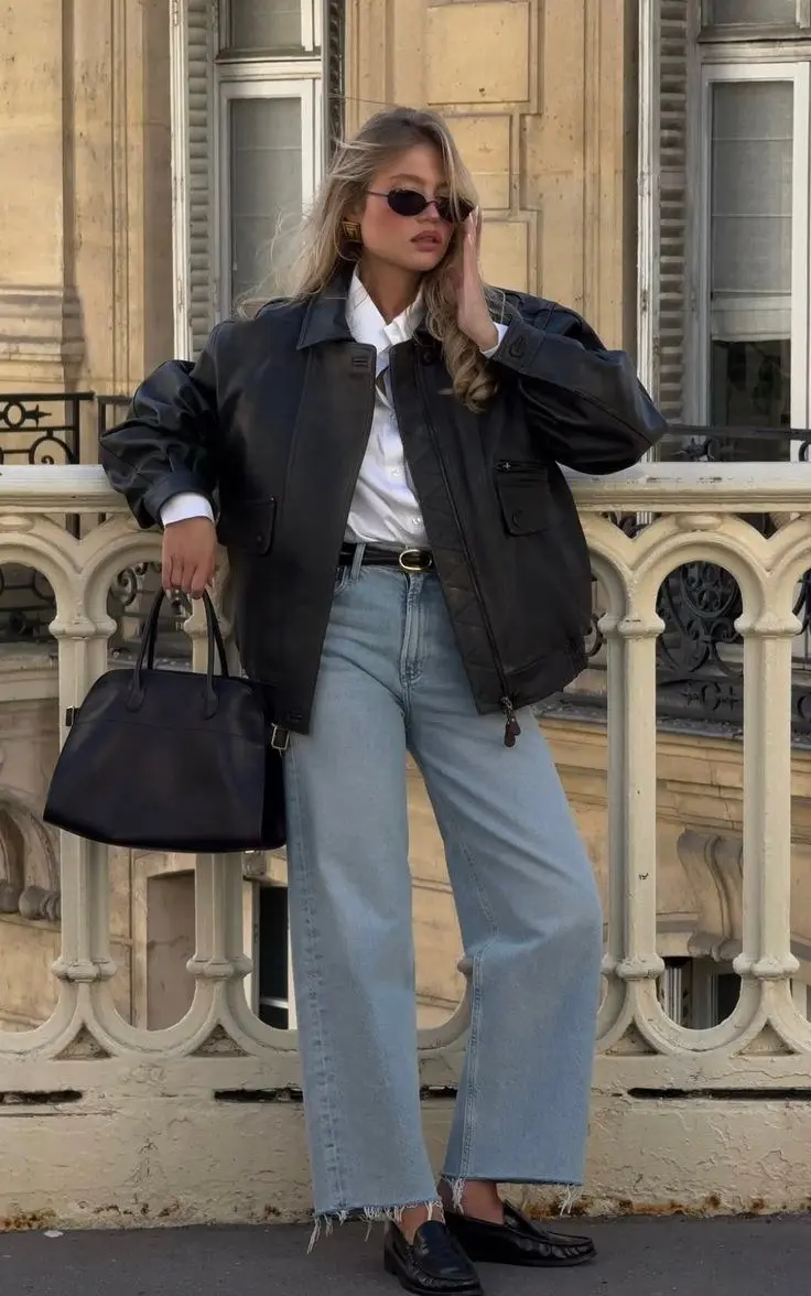 woman in black leather bomber jacket white shirt light blue wide leg jeans black belt black loafers large black structured tote small oval sunglasses gold hoop earrings balcony outdoor outfit 15