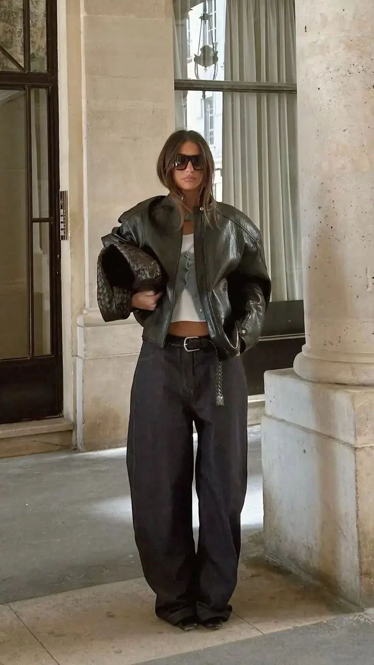 woman in dark olive green oversized leather bomber jacket white crop top dark charcoal barrel jeans leather belt large dark woven bag oversized sunglasses outdoor architecture outfit 19
