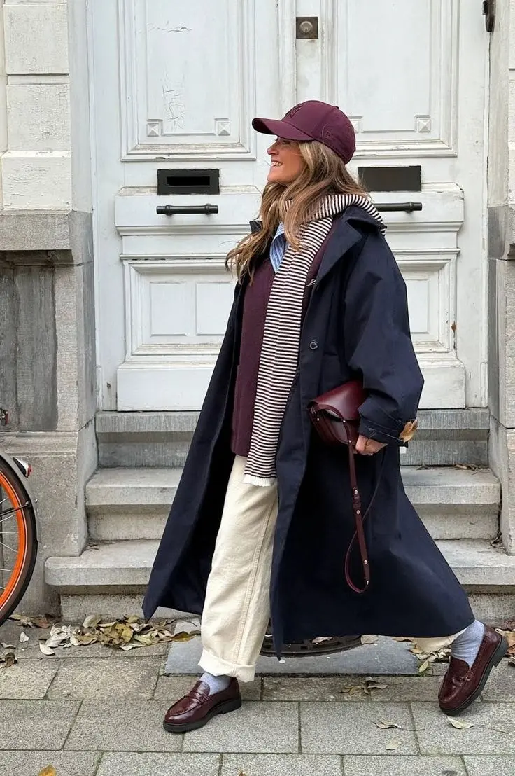 woman wearing navy long trench coat over burgundy vest striped scarf blue shirt cream trousers burgundy loafers and burgundy cap outfit 1