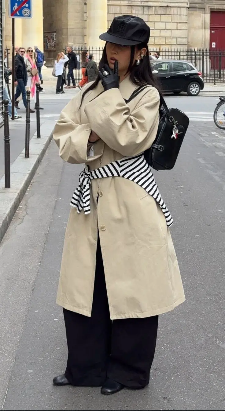 woman wearing beige long trench coat with black and white striped sweater tied at waist black wide leg trousers black leather gloves black backpack black flat boots black baker boy cap outfit 5