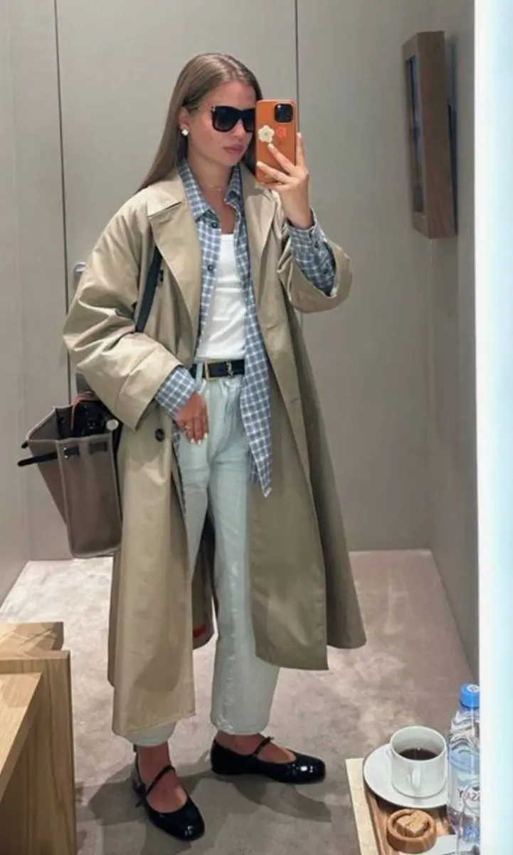 woman in mirror selfie wearing camel long trench coat open over red gingham shirt white tee black straight trousers black mary jane flats vintage canvas bag outfit 8