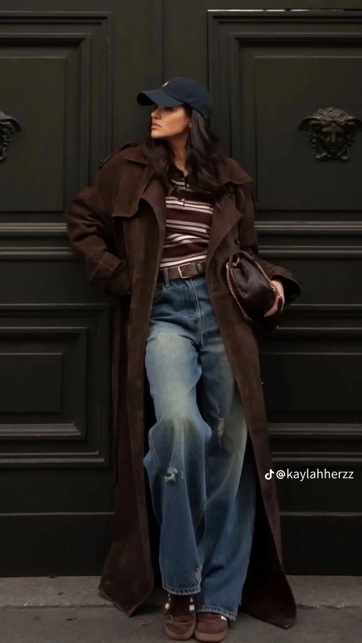 woman wearing dark brown suede long trench coat open over burgundy white striped polo wide leg blue jeans dark brown chain bag navy baseball cap dark brown sneakers outfit 9