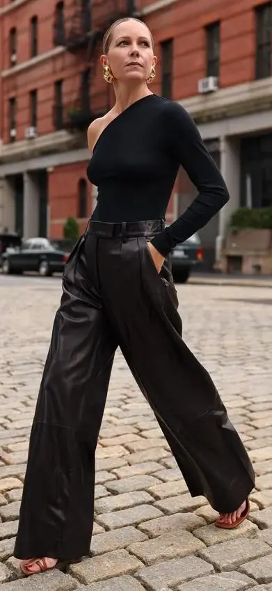 woman outdoors on cobblestones wearing black one-shoulder bodysuit, wide leg black leather trousers, terracotta strappy sandals, gold hoop earrings, outfit 3