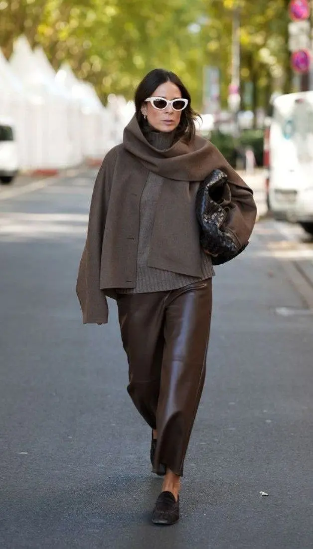 woman walking on street in full brown tonal outfit: chunky knit turtleneck, brown wool oversized cape, brown leather pants, dark loafers, white cat-eye sunglasses, holding woven dark bag, outfit 4
