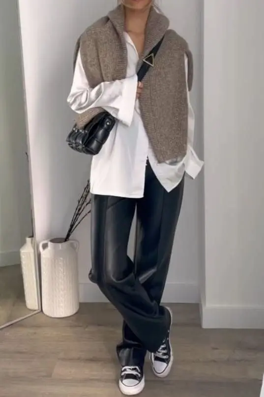 person wearing camel-tan collared knit sweater vest over oversized white shirt, wide leg black faux leather pants, black and white high-top Converse sneakers, small black crossbody bag with triangle hardware, outfit 8