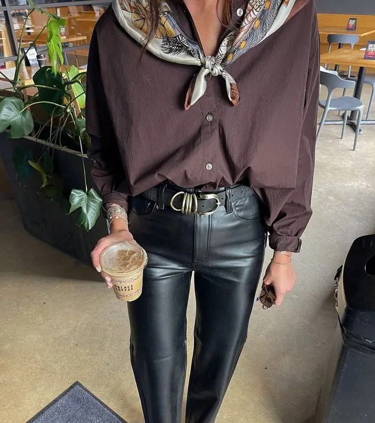 woman holding iced coffee wearing dark brown button-down shirt, printed silk scarf tied at neck, black straight-leg leather pants, wide black leather belt with gold buckle, bracelets, outfit 9