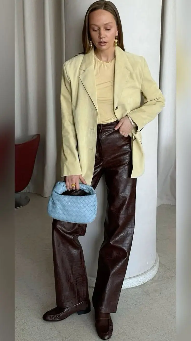woman indoors wearing butter yellow oversized blazer, pale yellow sheer top, chocolate brown croc-embossed wide leg leather pants, brown flat loafers, blue woven handbag, gold drop earrings, outfit 10