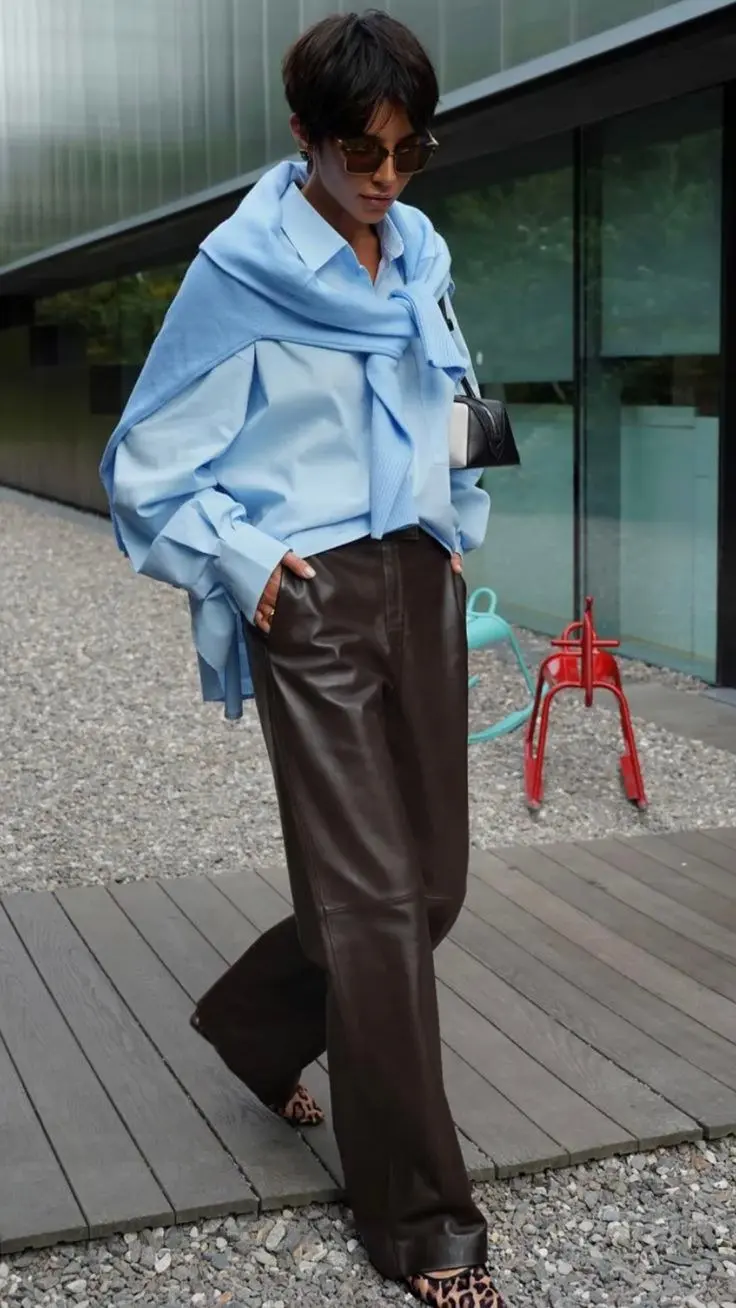 woman outdoors wearing baby blue oxford shirt, white underlayer, camel cashmere scarf draped over shoulders, dark chocolate wide leg leather pants, leopard print heeled mules, brown sunglasses, small black structured bag, outfit 12