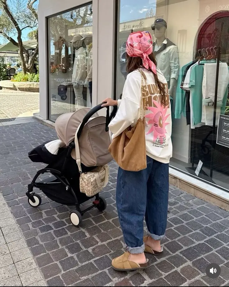 woman from behind pushing stroller in white graphic hoodie rolled blue jeans pink head scarf camel tote bag UGG slides outfit 2