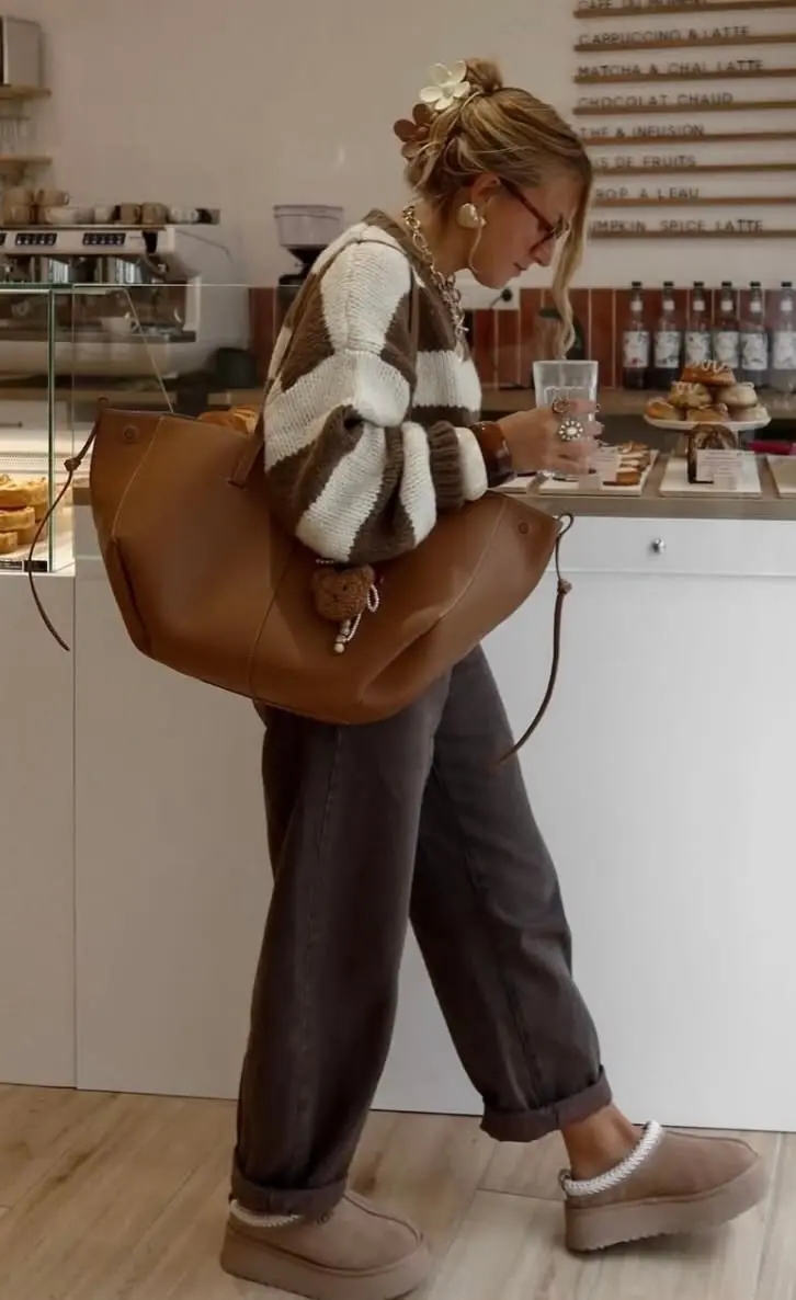 woman in cafe holding drink in brown and cream chunky striped sweater dark grey barrel trousers tan leather tote platform UGG boots pearl earrings floral hair clip glasses outfit 8