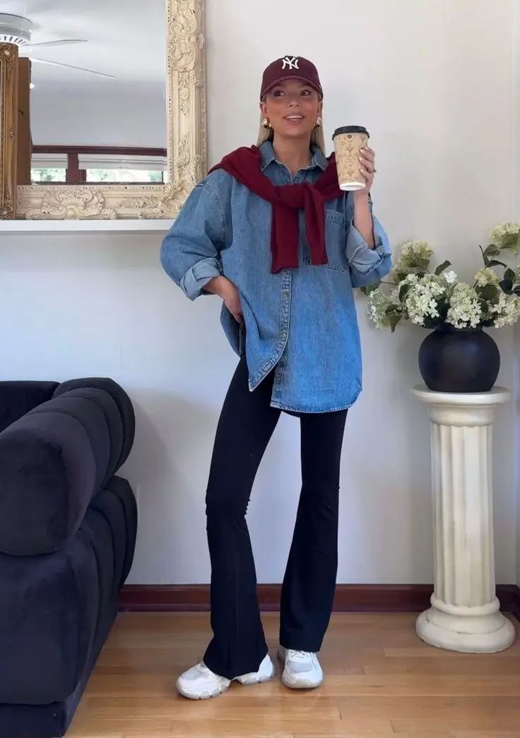 woman in oversized denim shirt as dress over black flare leggings burgundy knit tied on shoulders white sneakers maroon cap pearl studs coffee cup indoor outfit 11