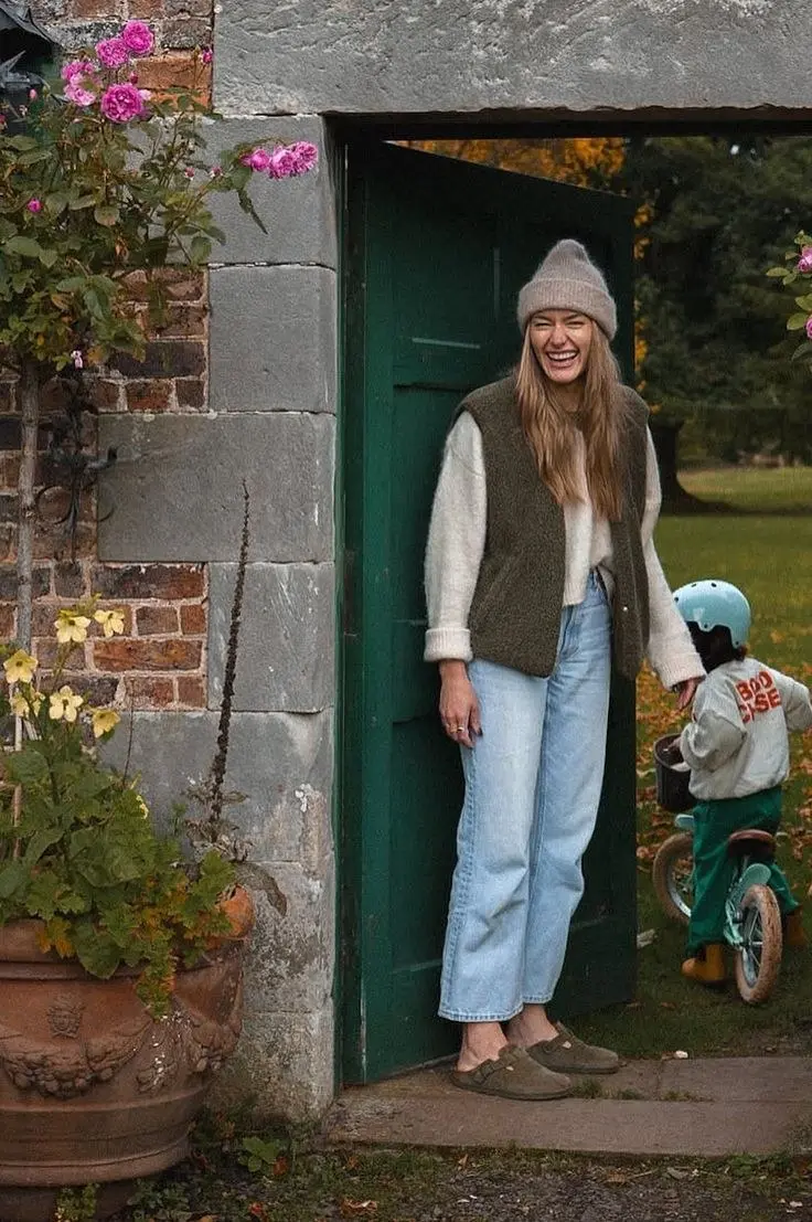 woman laughing in doorway in olive sherpa vest cream knit sweater straight light wash jeans mocha beanie olive suede clogs outdoor garden outfit 13