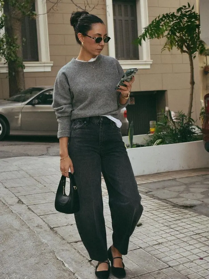 woman on street holding phone in grey crewneck sweater white tee dark barrel jeans black Mary Jane flats small black gold clasp bag sunglasses hair bun outfit 16