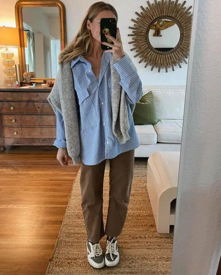 woman mirror selfie in blue striped oversized oxford shirt brown wide trousers grey knit cardigan draped on shoulders retro sneakers indoor outfit 23